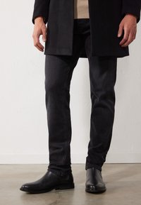 Botas de cuero negro, jeans oscuros con una sutil textura y un abrigo oscuro sobre un suéter claro. Enfoque en la parte inferior del cuerpo y el calzado.