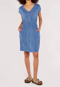 Robe en coton bleue avec manches courtes, col en V et taille avec cordon de serrage. Dotée d'un motif à pois blanc et de poches. Associée à des sandales beige.