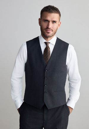 Mand med kort brunt hår og skæg iført en hvid skjorte, mønstret slips og mørkegrå knappet vest, stående med hænderne i lommerne.