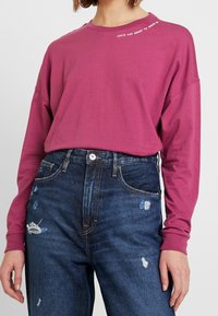 Langarmshirt aus magentanem Baumwollstoff mit Rundhalsausschnitt und weißen Textdetails an der Schulter. Getragen mit verspielten dunkelblauen Jeans.