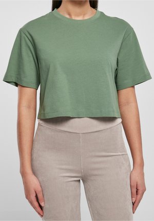 Kobieta ubrana w krótki, zielony t-shirt w kolorze szałwii z krótkimi rękawami oraz beżowe, prążkowane spodnie z wysokim stanem, stoi z ramionami luźno opuszczonymi wzdłuż ciała.