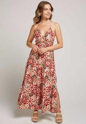 Robe maxi à motif floral rouge, noir et crème. Présente un décolleté halter, un détail en trou de serrure et des fentes sur les côtés. Portée avec des sandales compensées beiges.
