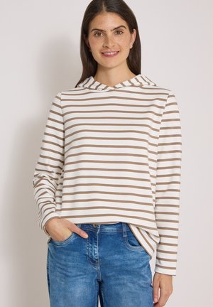 Kvinde smiler iført en hvid hættetrøje med lange ærmer med lysebrune horisontale striber og blå jeans, med den ene hånd i lommen.