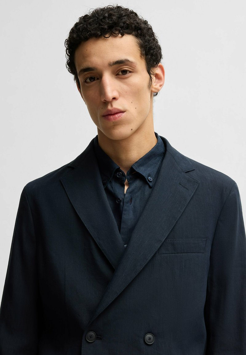 Marineblauer, doppelt geknöpfter Blazer aus strukturiertem Stoff, mit einer einzelnen Brusttasche und Reverskragen, getragen über einem dunklen Hemd.