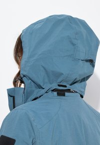 Chaqueta impermeable azul con capucha, cordones ajustables, tejido texturizado y un parche negro en el hombro para un detalle adicional.