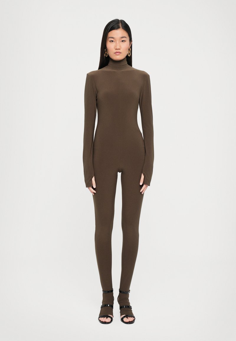 Brown long-sleeve jumpsuit with a turtleneck design, form-fitting shape, and side cutouts, paired with black open-toe sandals.