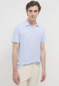 Jeune homme avec des cheveux bouclés courts portant un polo à manches courtes bleu clair et un pantalon blanc cassé, debout devant un fond uni.