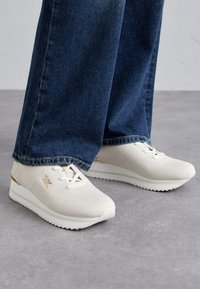 White textured sneakers with gold accents, featuring a lace-up design and thick white soles, paired with dark blue denim jeans.