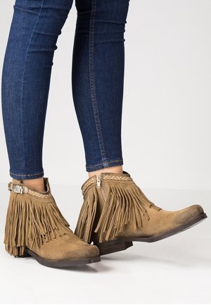 Bottes chelsea à franges en daim tan avec fermeture éclair sur le côté, détail en tresse décoratif et talon plat. Portées avec un jean skinny bleu foncé.