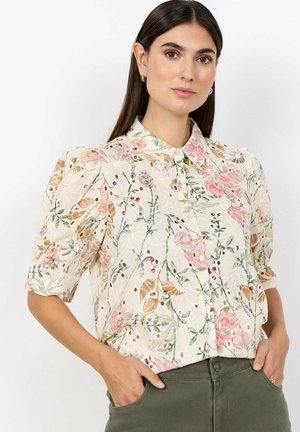 Vrouw draagt een lichte bloemenblouse met pofmouwen en een puntige kraag, gecombineerd met hooggetailleerde groene broek, handen in de zakken.