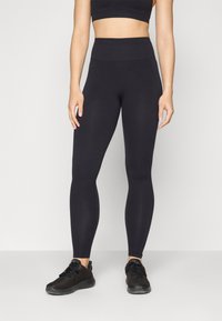 Svart høyhælte leggings i et glatt, elastisk stoff, kombinert med svarte joggesko. Designet er tettsittende og sømløst.