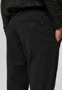 Detailní záběr černých kalhot s zadní kapsou na knoflík a malou visačkou Esprit, nošených s tmavým košilem s vzorem a rukou v přední kapse.