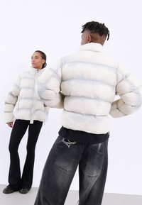 Puffjacka i krämfärg och grått med dragkedja, som har hög krage och quiltade sektioner. Bärs med svarta utsvängda byxor och slitna jeans.
