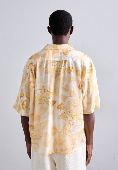 Chemise à manches courtes en tissu jaune clair au toucher doux, ornée de motifs circulaires orange et de texte imprimé. Modèle présenté de dos.
