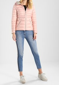 Jasnoróżowa kurtka puchowa z zamkiem błyskawicznym, z kapturem i bocznymi kieszeniami. Nosi na sobie czarną bluzkę i jasnoniebieskie dżinsy z bocznymi paskami.