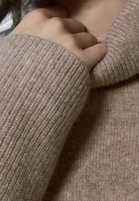 Dłoń chwytająca kołnierz miękkiego, prążkowanego beżowego swetra blisko szyi i ramienia.