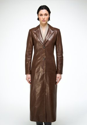 Woman with short dark hair wearing a long brown leather coat over a beige top, standing against a plain white background.