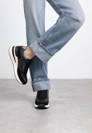 Zapatillas negras con un upper de cuero liso y acentos de ante, que cuentan con una suela blanca y punta redonda, combinadas con jeans azul claro arremangados.
