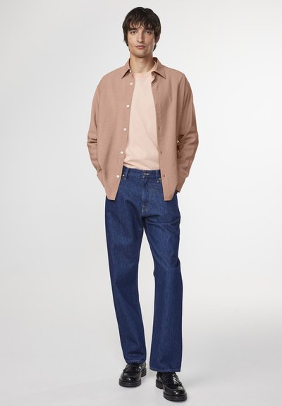 Camicia button-up marrone chiaro sopra una maglietta rosa chiaro, abbinata a jeans a gamba larga blu scuro e scarpe eleganti nere.