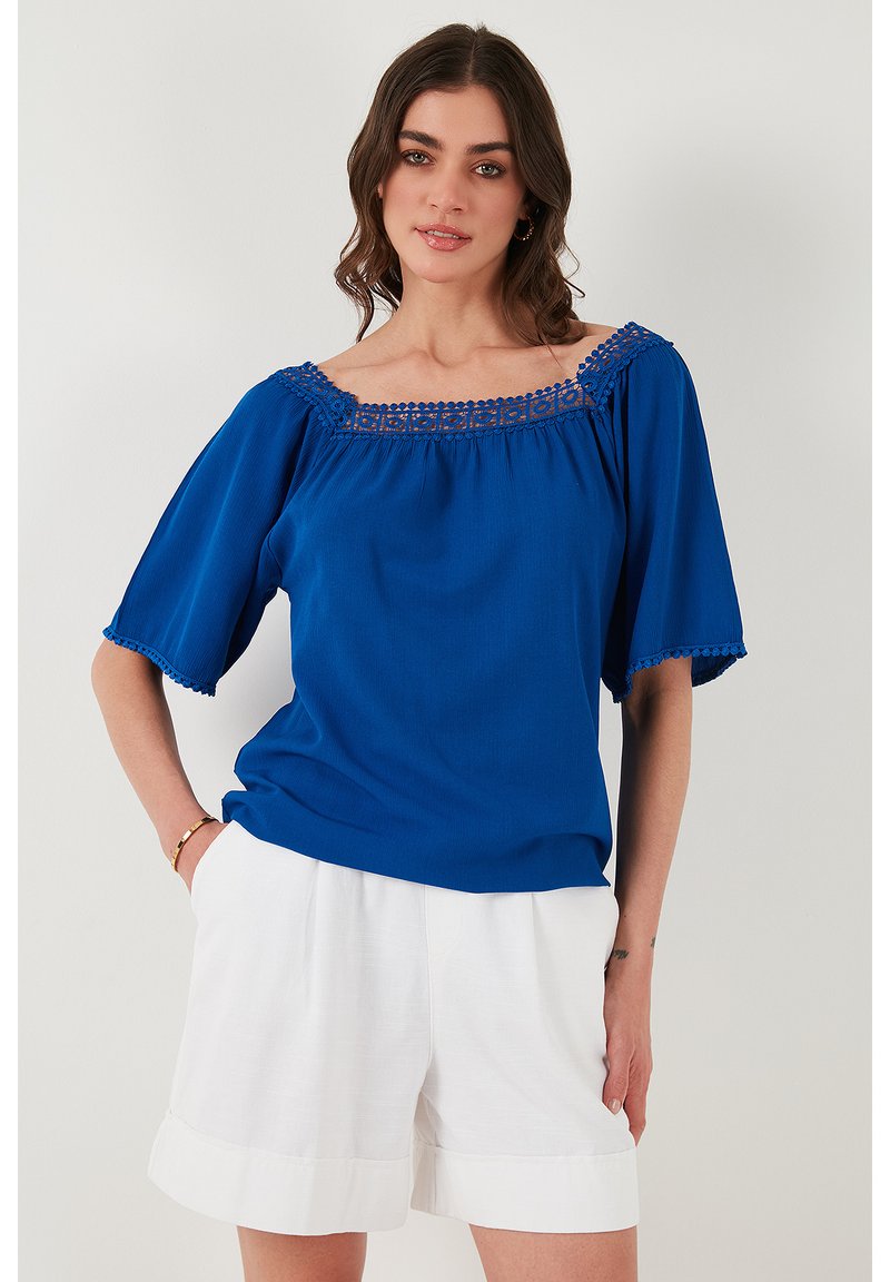 Modrá blúzka s off-shoulder výstrihom a výšivkou. Voľný strih s krátkymi rukávmi. V kombinácii s bielymi šortkami, predvádzajúc letný štýl.