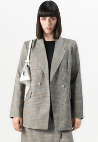 Grå rutete blazer med dobbeltspent design, spisse lapeller og sølvknappeebokser. Kombinert med en liten hvit håndveske.
