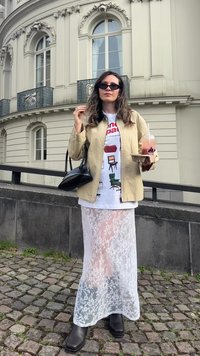 Gonna lunga in pizzo bianco, stivali neri a punta quadrata, giacca beige su una t-shirt grafica con illustrazioni di sedie. Tieni in mano una bevanda e una borsa piccola.