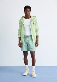 Mint green zip-up hoodie over white tank top, paired with light green shorts. Shoes are beige high-tops, white socks, and logo accents.