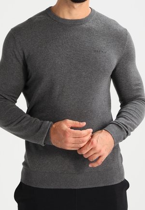Mann trägt eng anliegenden, dunkelgrauen Pullover mit langen Ärmeln, gerippten Bündchen und Saum, zieht mit beiden Händen die Ärmel hoch.
