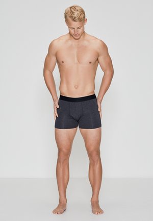 Sorte boxershorts lavet af strækbart stof, tætsiddende design og en blød elastisk talje. Modellen står barfodet med hænderne hvilende på lårene.