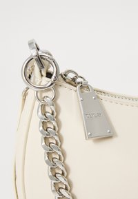 Sac à main crème avec une finition lisse, doté de quincaillerie en chaîne argentée et d'une étiquette logo. Comprend une fermeture éclair et des accents métalliques pour un design fonctionnel.