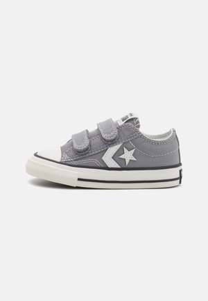 Zapatilla de lona gris con una punta de goma blanca, dos correas de Velcro, logo de estrella y una franja negra en la suela. Etiqueta de logo en la parte trasera visible.