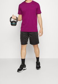 Fialové tričko Nike, černé šortky, držící kettlebell. Černé sportovní boty se šedými akcenty a černé ponožky. Jednoduchý design sportovního oblečení.