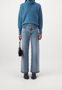 Pull à col roulé bleu tricoté, jean large taille haute bleu clair avec ourlet effiloché, et sac à main noir. Chaussures noires épaisses.