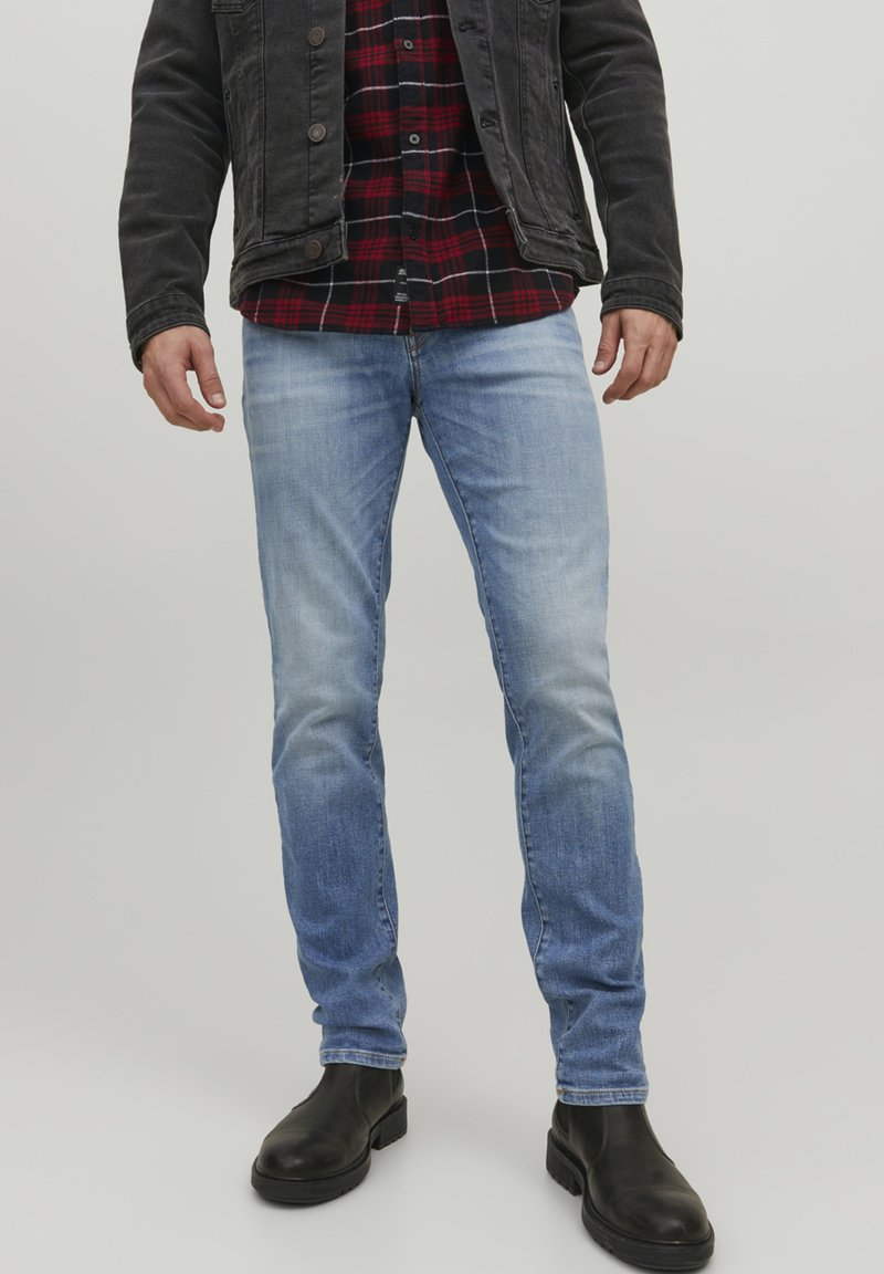 Jeans de denim de culoare albastru deschis, cu un finisaj ușor decolorat, asortați cu o jachetă neagră și o cămașă cu carouri roșii. Bocanci negri la gleznă completează ținuta.