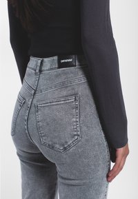 Persona vistiendo jeans gris de cintura alta con la etiqueta DRDENIM, combinados con una blusa ajustada de manga larga negra, mostrada desde la espalda.