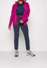 Fleece bunda v jasně růžové barvě, doplněná námořnickým tričkem a úzkými námořnickými kalhotami. Obuv má béžové a modré akcenty, vhodná pro venkovní použití.