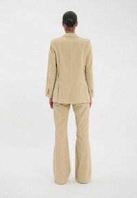 Costume en velours côtelé beige comprenant un blazer ajusté et un pantalon évasé. Les détails notables incluent des accents de boutons sur les manches et une coupe structurée.