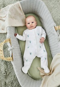 Weißer Baby-Body mit floraler Stickerei, auf einem grünen Stoff in einem geflochtenen grauen Kinderbett mit strukturierten Kanten ausgelegt. Weiche, gemütliche Decken sind in der Nähe.