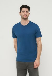 Άνδρας με σκούρα μαλλιά και γενειάδα, φοράει απλό μπλε μπλουζάκι και σκούρο γκρι τζιν, στέκεται μπροστά σε έναν απλό υπόλευκο φόντο.