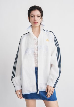 Ženska oblečena v belo Adidas trenirko s črnimi črtami in modro jeans krilo, stoji ob preprostem svetlem ozadju.