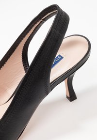 Escarpin à talon haut en cuir noir à bride arrière avec semelle intérieure beige et couture visible sur les lanières, présenté en angle sur fond blanc.