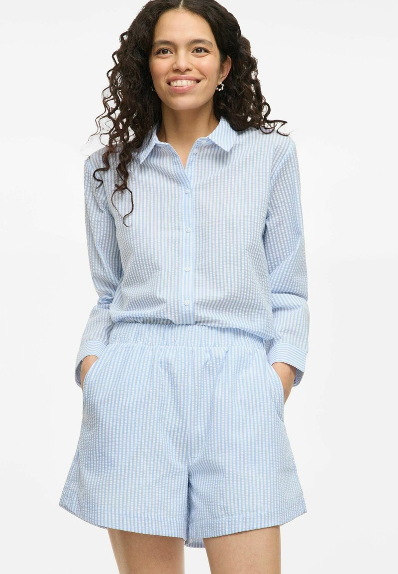 Chemise bleue clair à boutons avec des rayures blanches verticales, associée à un short à taille élastique assorti, comportant des poches latérales.