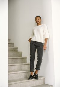 Una donna è in piedi su scale di cemento indossando un maglione bianco e peloso, jeans neri decorati con perle, mocassini neri, orecchini a cerchio e un braccialetto.
