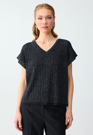 Kobieta o jasnych włosach, ubrana w czarną prążkowaną bluzkę z krótkim rękawem i dekoltem w serek oraz czarne spodnie, stojąca na jednolitym jasnym tle.