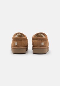 Καφέ σουέτ πάνινα παπούτσια slip-on με μαλακή επένδυση, στρογγυλεμένες άκρες και επίπεδες καουτσούκ σόλες. Μικρή ετικέτα μάρκας στη φτέρνα. Μινιμαλιστικός σχεδιασμός.