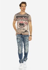 Beige grafische t-shirt met rode en zwarte letters, gecombineerd met versleten blauwe denim jeans met ingewikkelde stiksels. Witte sneakers.
