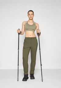Olivinės žygių kelnės ir derantis sportinis liemenukas. Kelnės turi užtrauktukais užsegamas kišenes, o modelis laiko žygių lazdas. Juodi žygių batai.