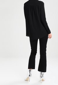 Persona che indossa una blusa nera larga, pantaloni neri aderenti con orli svasati e stivaletti alti con tacco a spillo argentati, in piedi con le spalle rivolte verso la fotocamera.