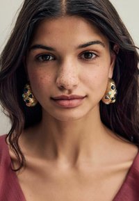 Goldene Creolenohrringe mit bunten Edelsteinen in verschiedenen Formen. Das Model hat lange dunkle Haare und trägt ein tiefes, kastanienbraunes Oberteil.