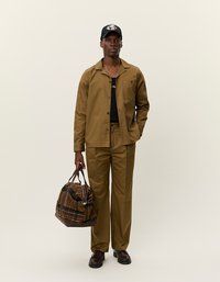 Barna gombos ing és ahhoz illő nadrág, alatta fekete ujjatlan felső. Barna kockás duffel táskát hordoz, és fekete sapkát visel.